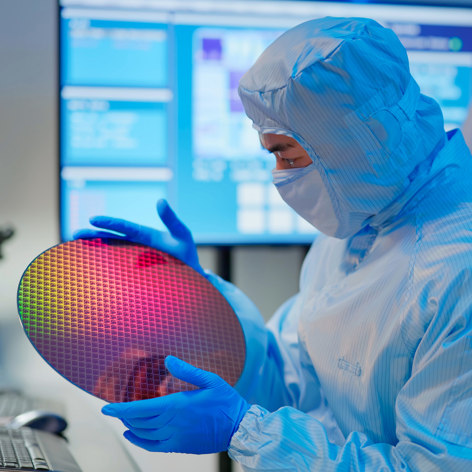 Male technician in sterile coverall holds wafer that reflects many different colors with gloves and check it at semiconductor manufacturing plant.