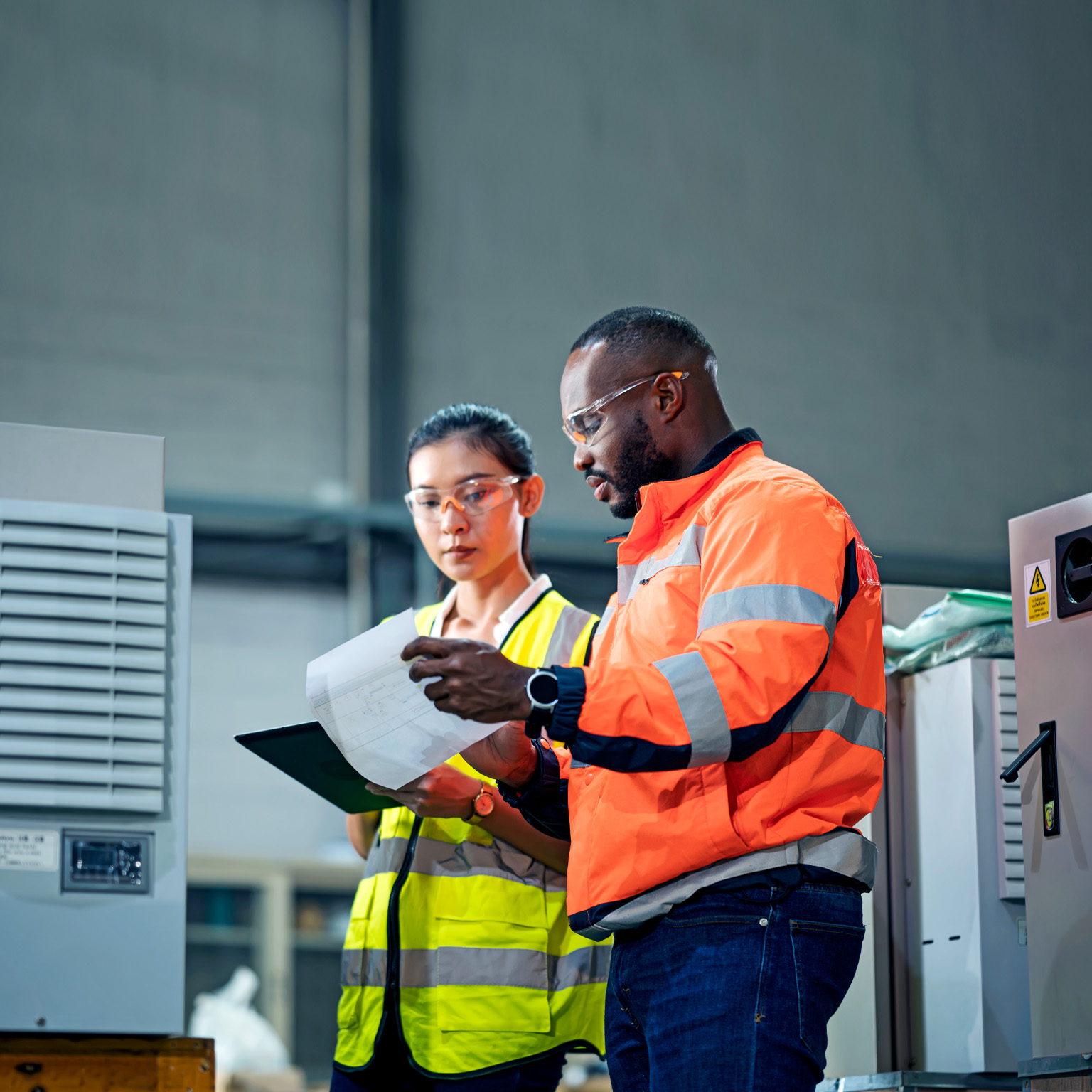 A woman holding a tab and a man with a checklist, both wearing different color-coded work jackets standing in a control room