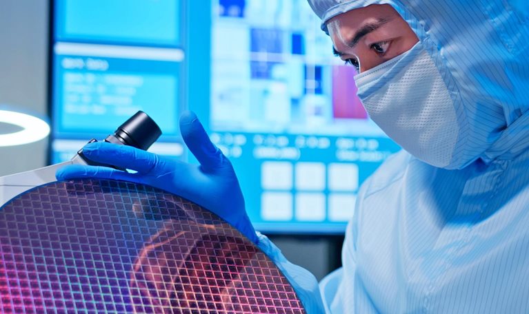 Asian male technician in sterile coverall holds wafer that reflects many different colors with gloves and check it at semiconductor manufacturing plant.