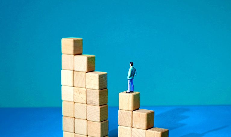 A series of wooden blocks arranged in a stair-like formation, with a gap in the middle where a vertical set of blocks is missing. A tiny man stands, unable to continue forward with his arms hanging at his sides.
