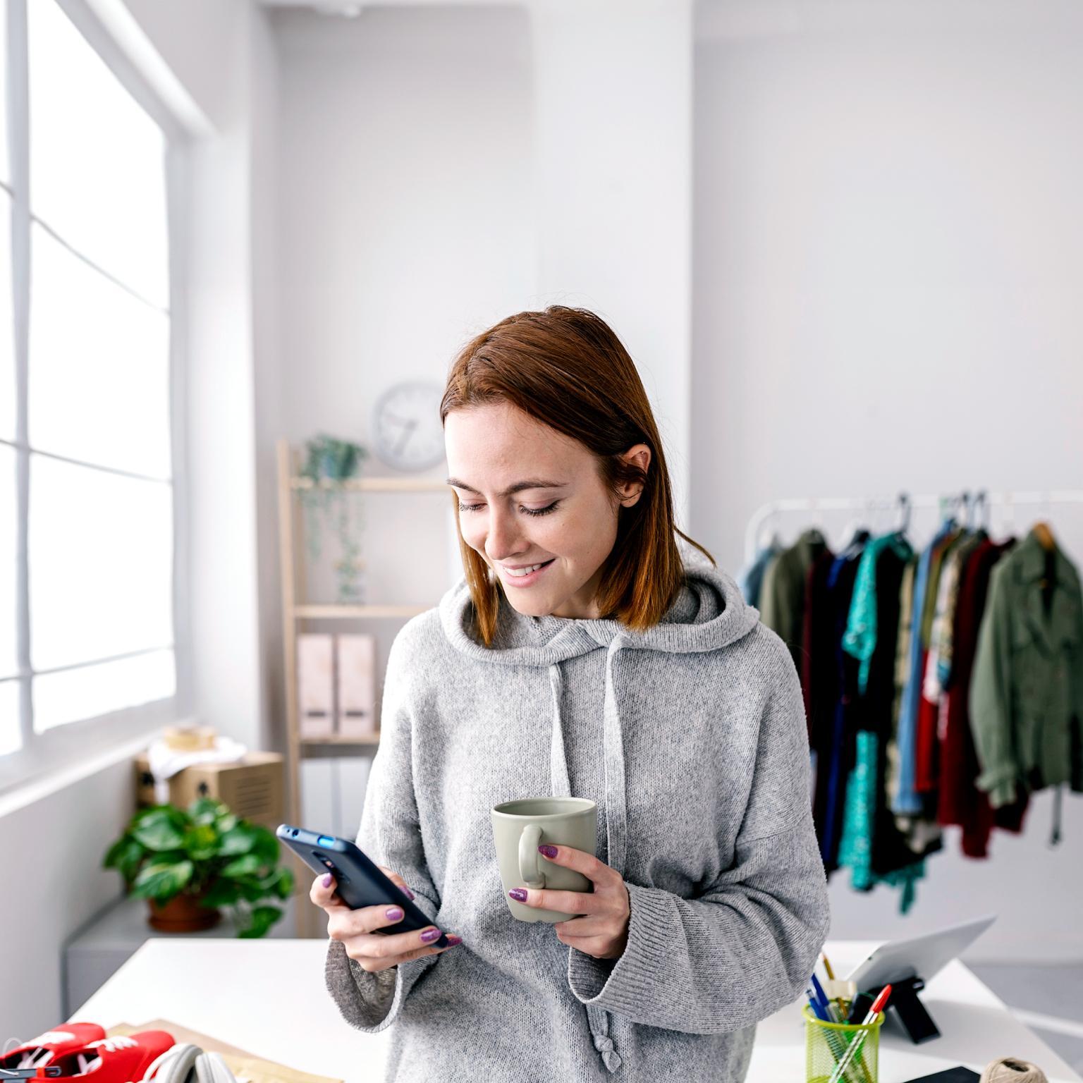 Woman dressed in gray hoodie looking down and phone in one hand, other hand holding ceramic coffee mug.
