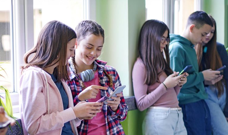 A group of teenagers are standing against a wall, looking at their smartphones. One of the girls in the middle is pointing at her phone, sharing something with another girl beside her.
