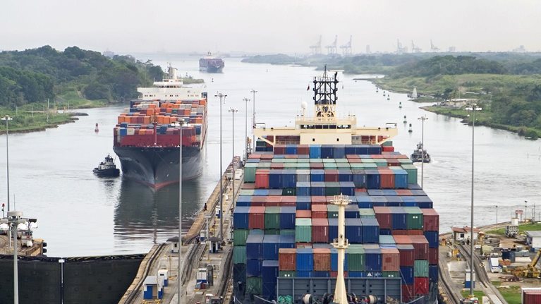 Cargo ships assisted by tugboats, entering the Panama Canal at Gatun Locks on the Atlantic side. The ships are fully loaded with cargo heading west towards the Pacific.