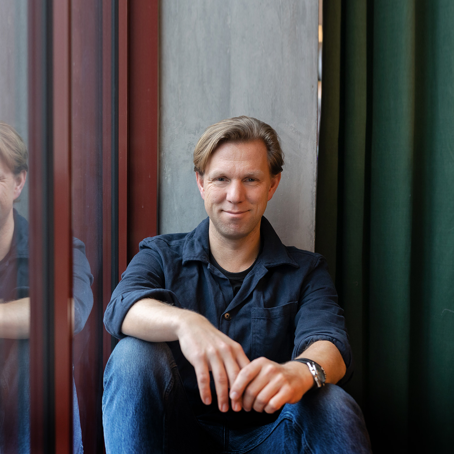 Portrait of Pelle Sommansson in jeans and a collared shirt. Sommansson has a relaxed pose and is leaning against an office window.