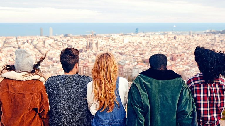 Image from behind of a group of friends sitting on a terrace looking out over a city with the ocean in the far distance.