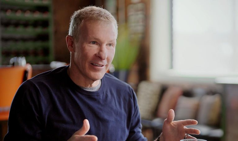 A middle-aged man gesturing while speaking at a table with mugs and glasses in a cozy, well-lit room.