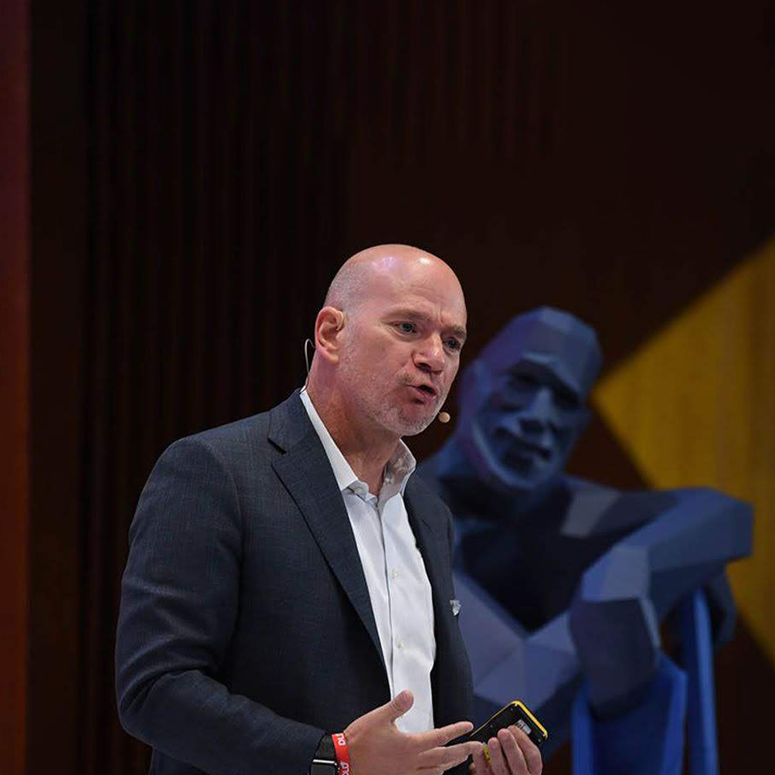  Andy McAfee speaking on stage with a smiling blue statue in a relaxed pose behind him.