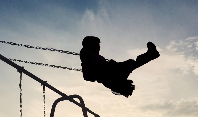 A silhouetted child soars through the air on a swing set. The setting sun casts a warm glow on the scene, creating a quiet and nostalgic atmosphere.