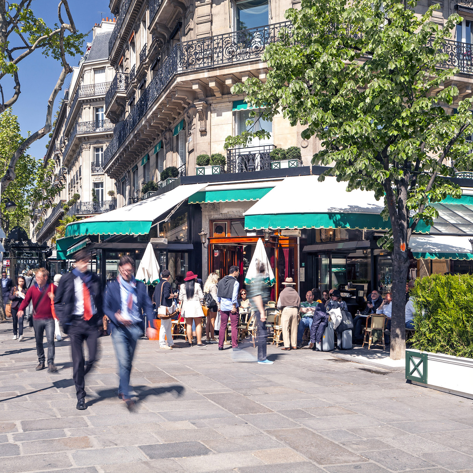 A busy Boulevard Saint-Germain in Paris. A corner café and sidewalk are full of professionals and people going about their day. Apartments are seen above the lower shops.