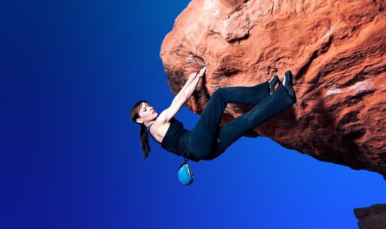 A woman in black rock-climbing clothing navigates a large overhanging bolder without a rope. She has a firm hold with both hands but seeks a foot hold as she makes her way over the protruding rock.