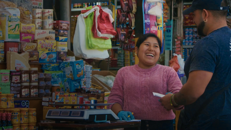 Photograph of a small grocery store checkout area showing a cashier wearing a pink sweater and gloves interacting with a customer in a blue shirt and cap. Shelves behind the cashier are stocked with various packaged food items, including biscuits, cereals, and snacks, highlighting a typical local market setting.