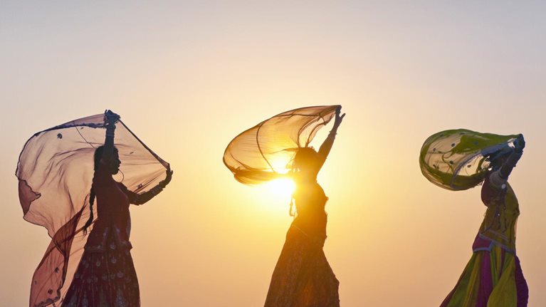 As the sun rises behind them, four Indian women dressed in traditional attire stroll along the crest of a desert dune, casting their figures in silhouette.
