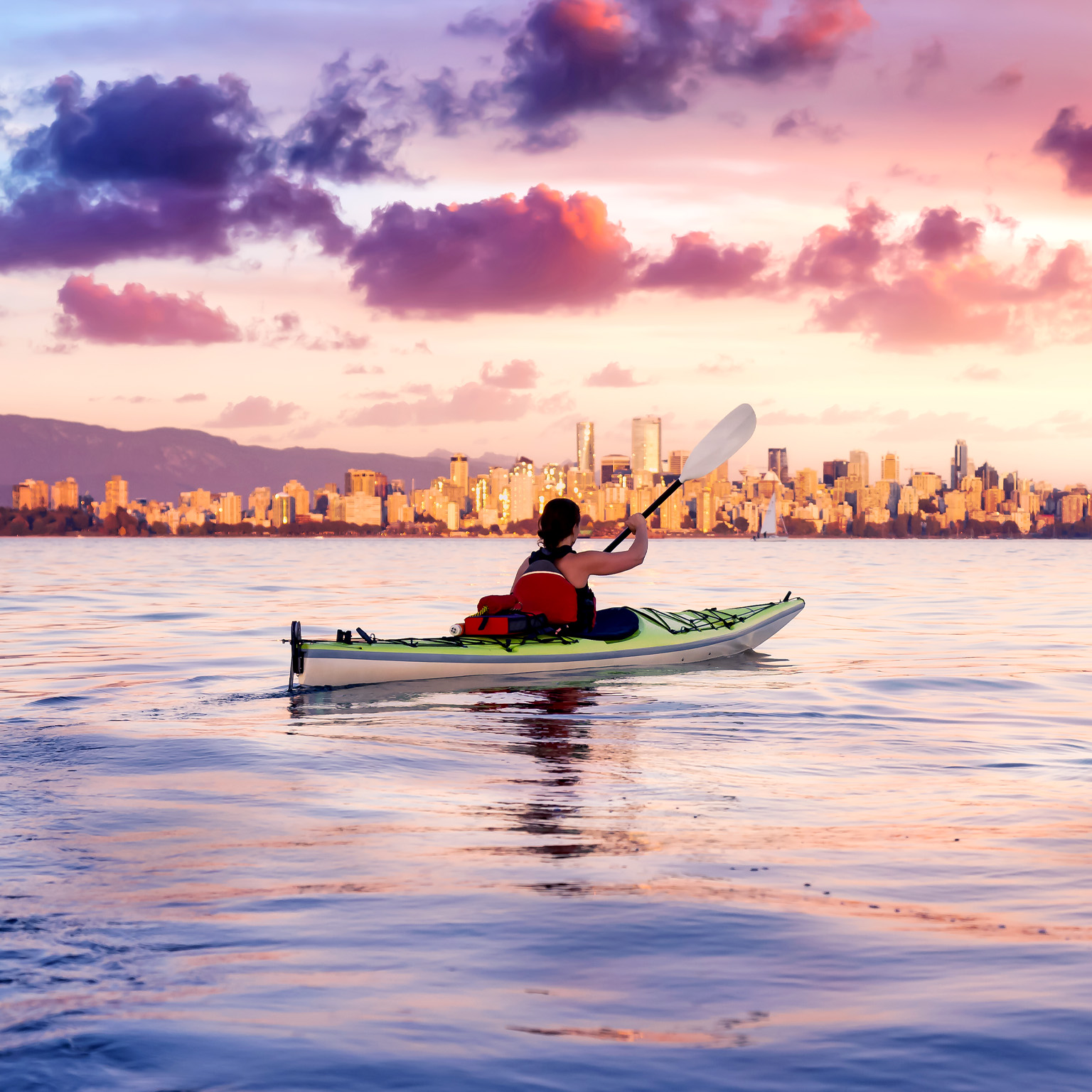 As the sun sets behind the Vancouver skyline, a woman paddles her kayak on a vast body of water. The vibrant sunset sky paints the horizon with shades of pink and purple, casting a mirror-like reflection on the water.