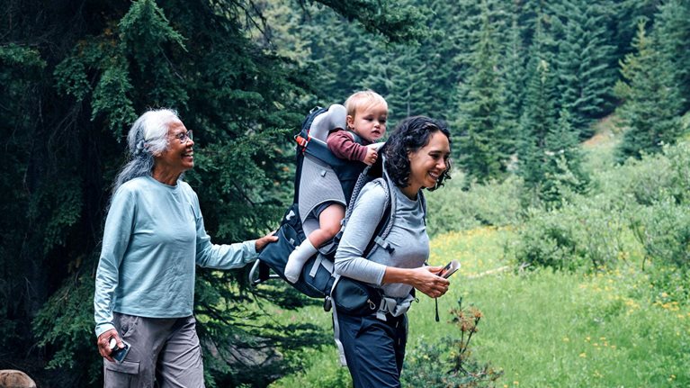A smiling family, composed of a grandmother, grandfather, mother with a baby in a carrier on her back, and a young girl, treks through a forest, crossing a wooden bridge over a small stream. The grandfather assists the girl as they walk.