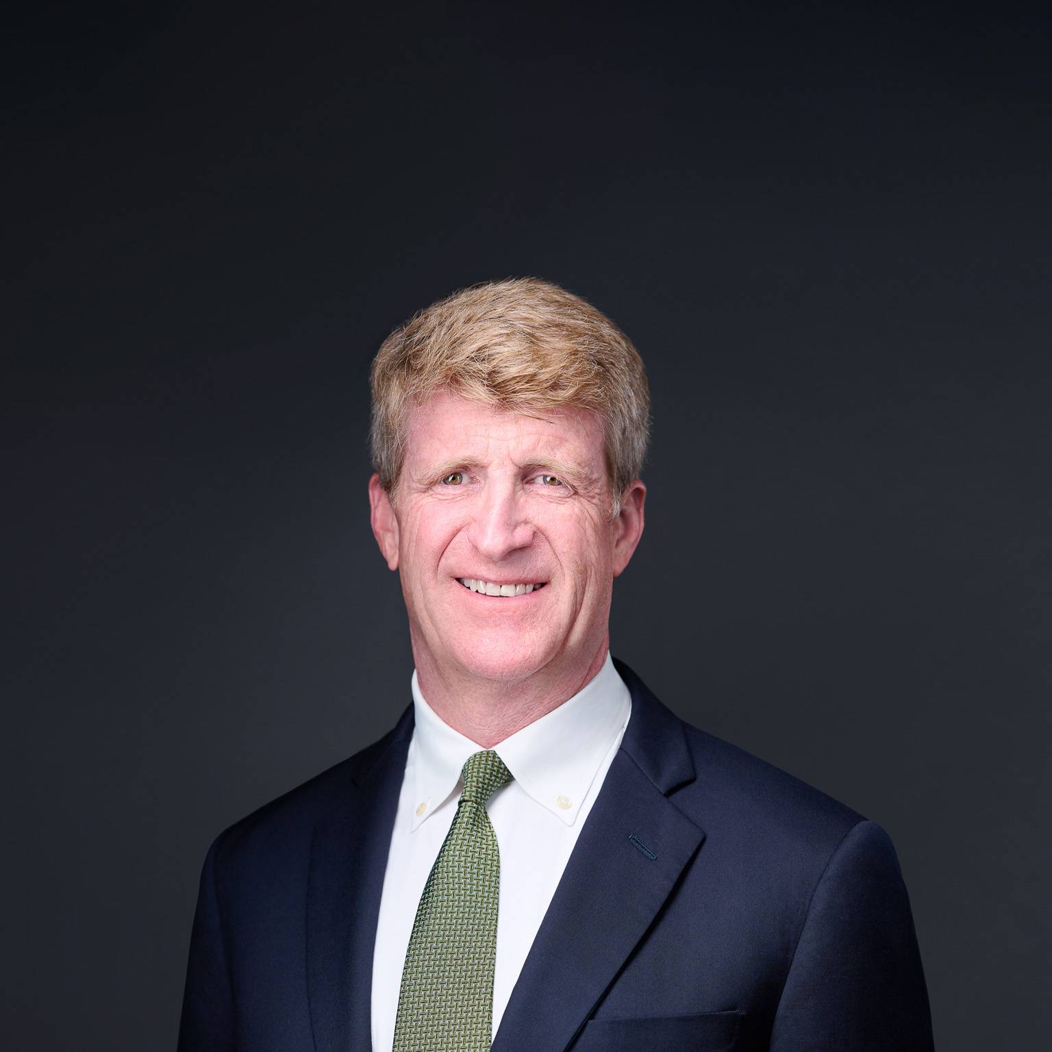Patrick J. Kennedy, wearing a navy suit jacket, a green tie, and a smile, photographed against a dark background.