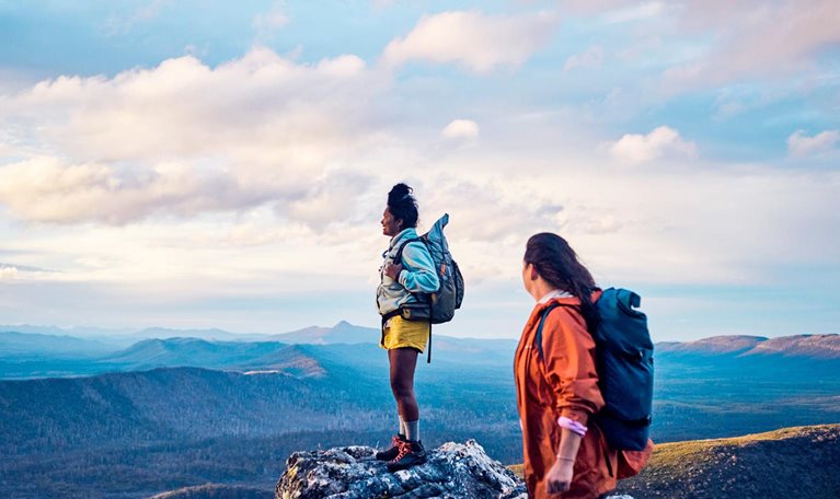 Two hikers stand on a rocky mountain peak, wearing backpacks and looking out over a vast, scenic landscape. The expansive view and soft light convey a sense of adventure, achievement, and connection with nature.