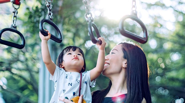 A mother smiles as she holds her young child up while the child grasps a monkey bar in an outdoor playground.