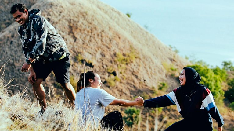 Three smiling friends, who are slightly heavyset, are climbing a grassy hill. One friend is assisting the other to stand up from a sitting position. The view from the top of the hill offers a peaceful ocean scene bathed in golden sunlight.