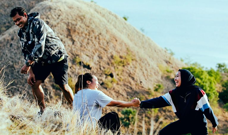 Three smiling friends, who are slightly heavyset, are climbing a grassy hill. One friend is assisting the other to stand up from a sitting position. The view from the top of the hill offers a peaceful ocean scene bathed in golden sunlight.