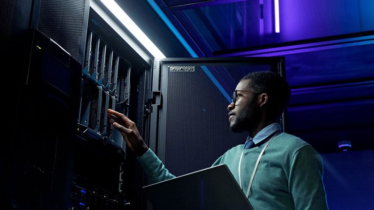 Low angle portrait of young African American data engineer working with supercomputer in server room lit by blue light and holding laptop