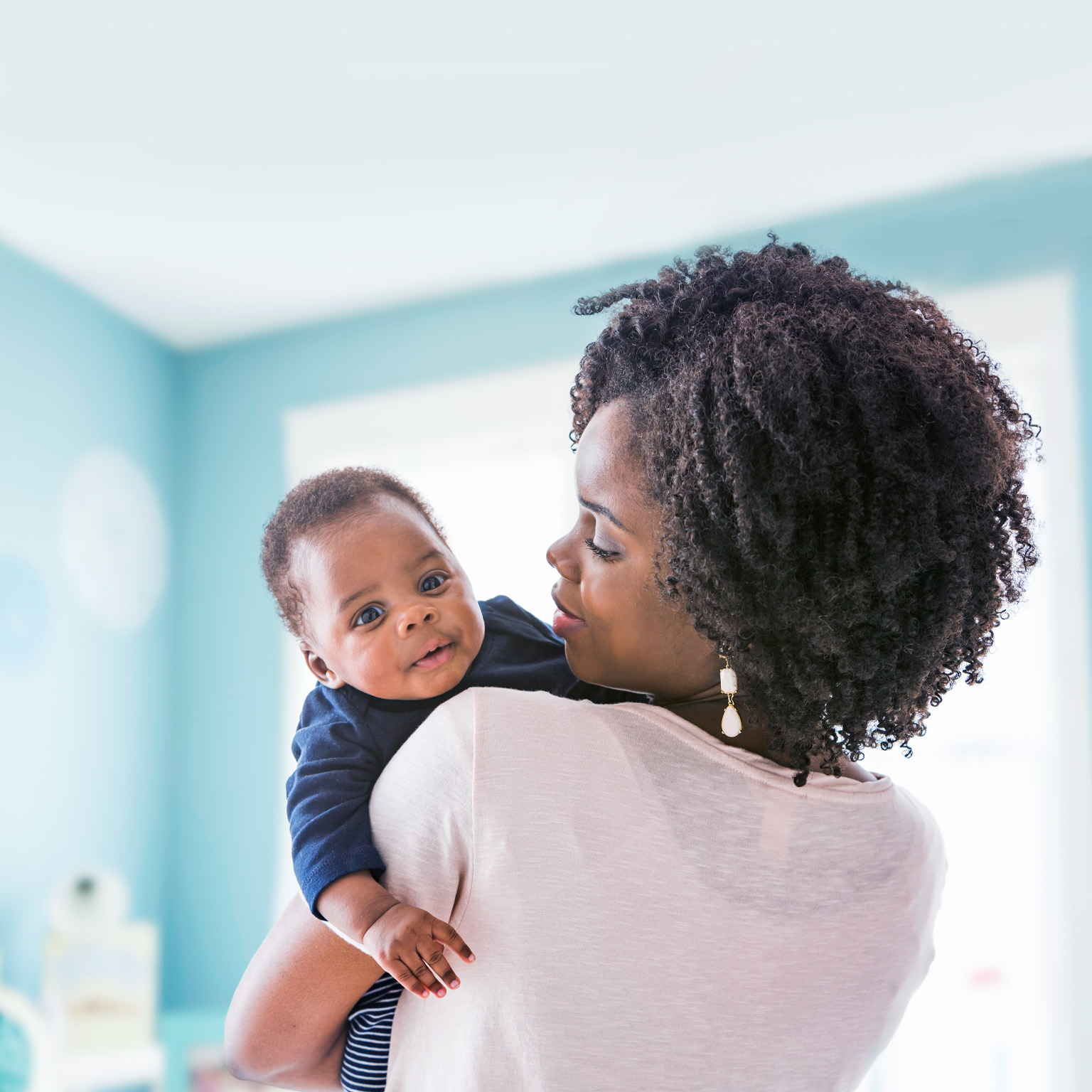 A Black mother with tightly curled ringlets holds her baby on her shoulder. The baby gazes at the viewer inquisitively as the mother looks at him thoughtfully.
