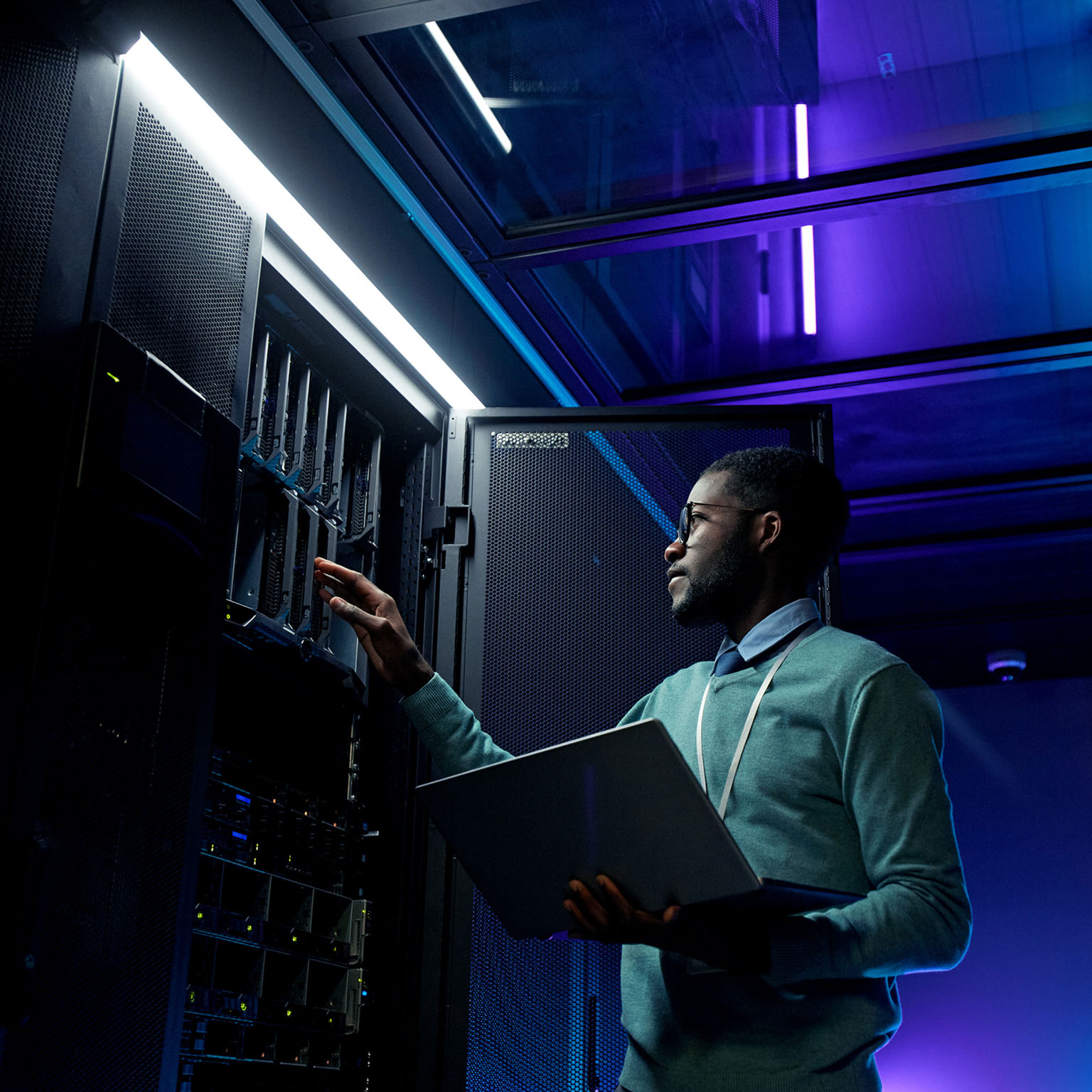 Low angle portrait of young African American data engineer working with supercomputer in server room lit by blue light and holding laptop