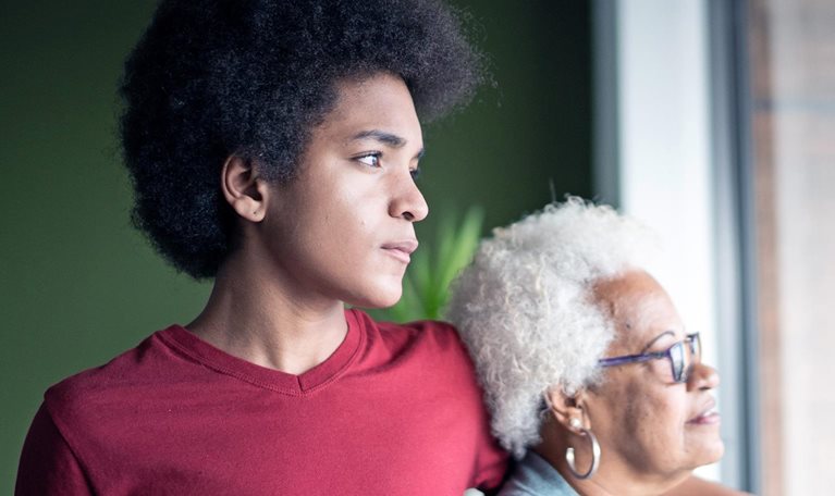 A young African American man stands beside his grandmother, his arm resting gently on her shoulder as they both gaze thoughtfully out of a window.